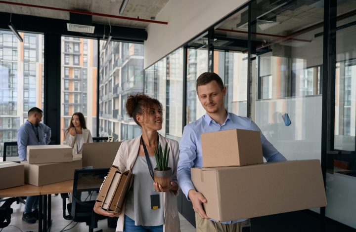 Corporate employee holding file folders and potted plant in hands and looking at colleague with cardboard boxes